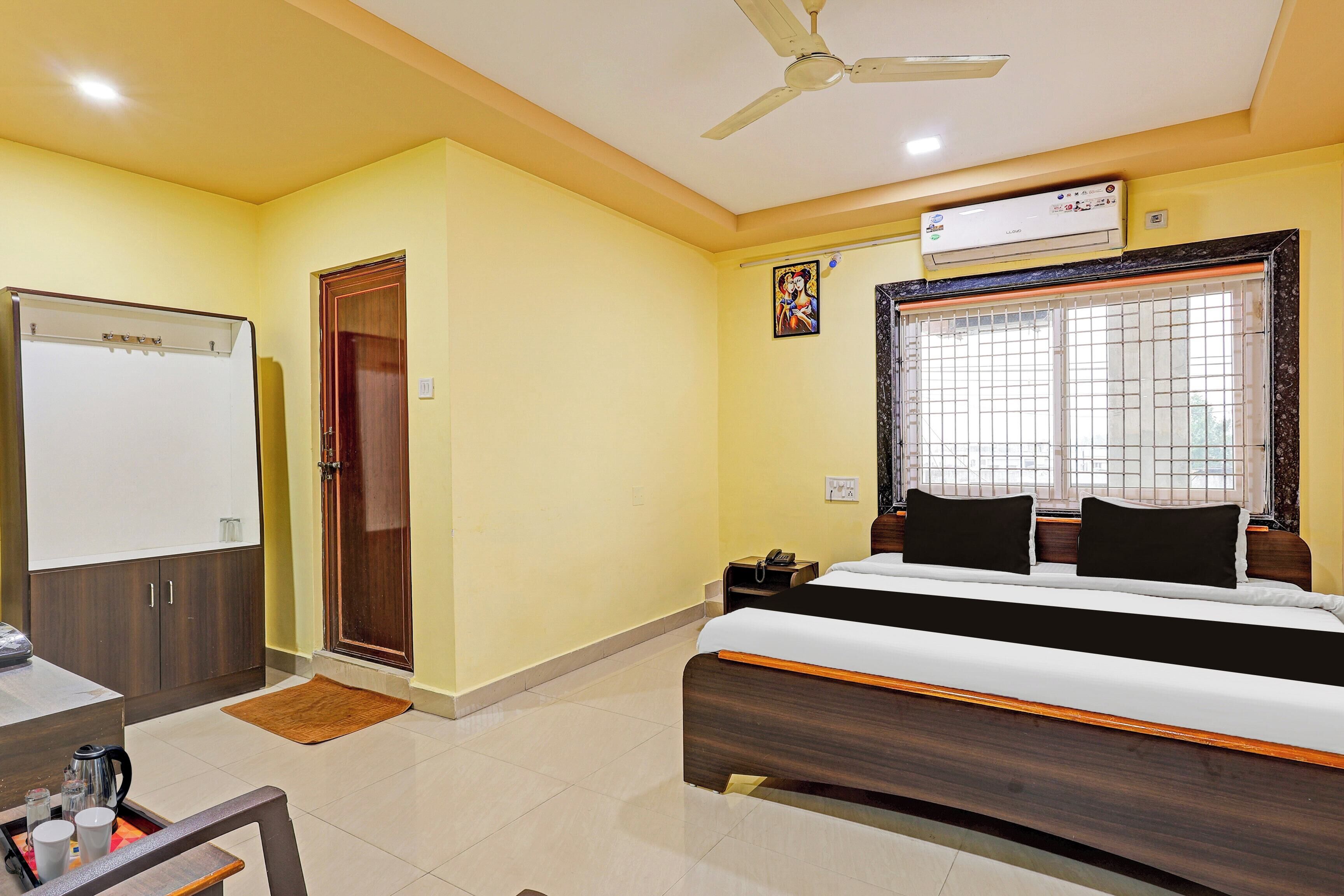A hotel room with a bed, a desk, a chair, a wardrobe, a door, a ceiling fan, a window with bars, and an air conditioning unit.