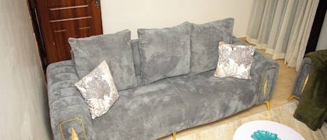 A grey sofa with patterned cushions, a small white table with a decorative item, and a wooden door in a room with a tiled floor.