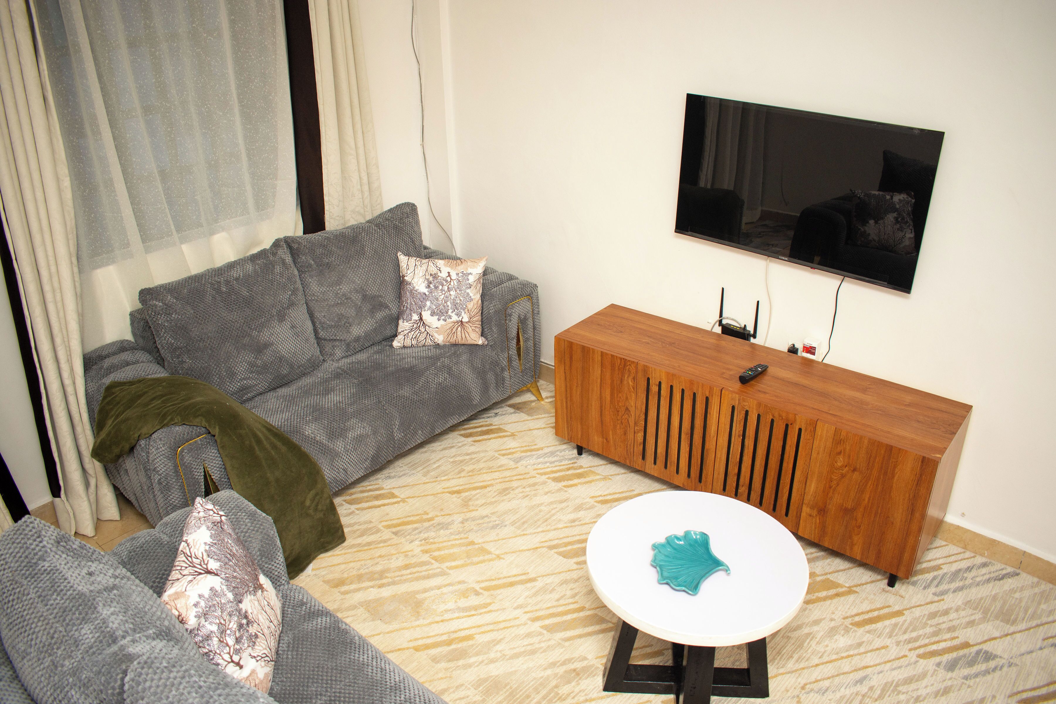 Ein Wohnzimmer mit grauem Sofa, einer Holzkommode, einem Flachbildfernseher und einem kleinen, weißen Tisch mit einem blaugrünen Gegenstand.