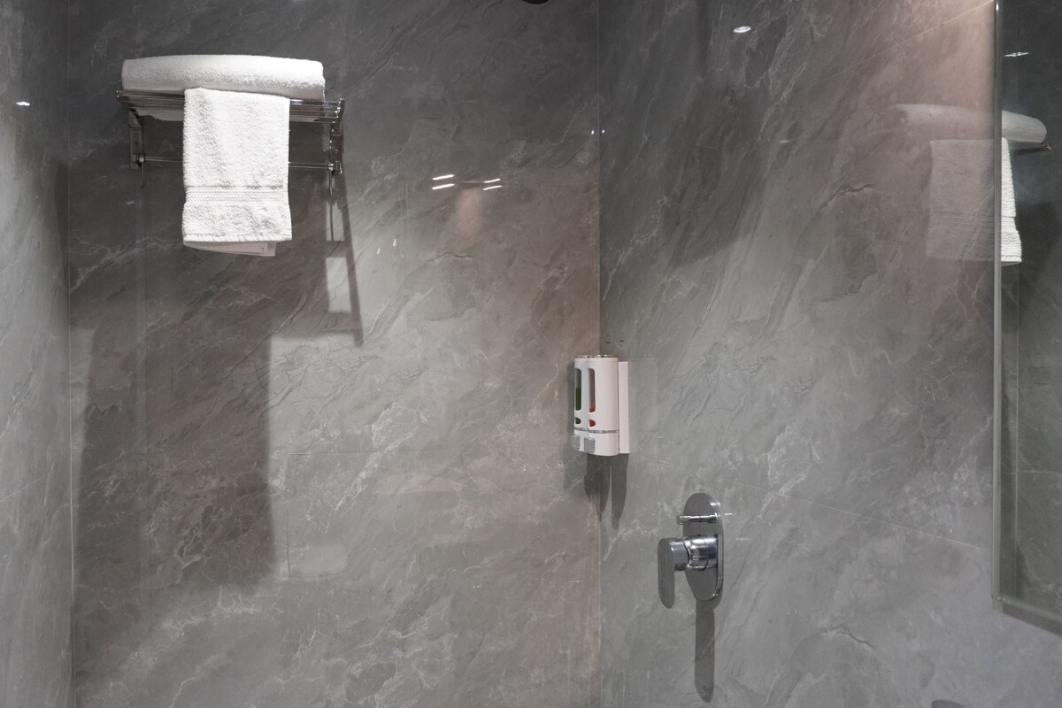 A modern bathroom with a glass shower enclosure, a white towel hanging on a rack, and a soap dispenser.