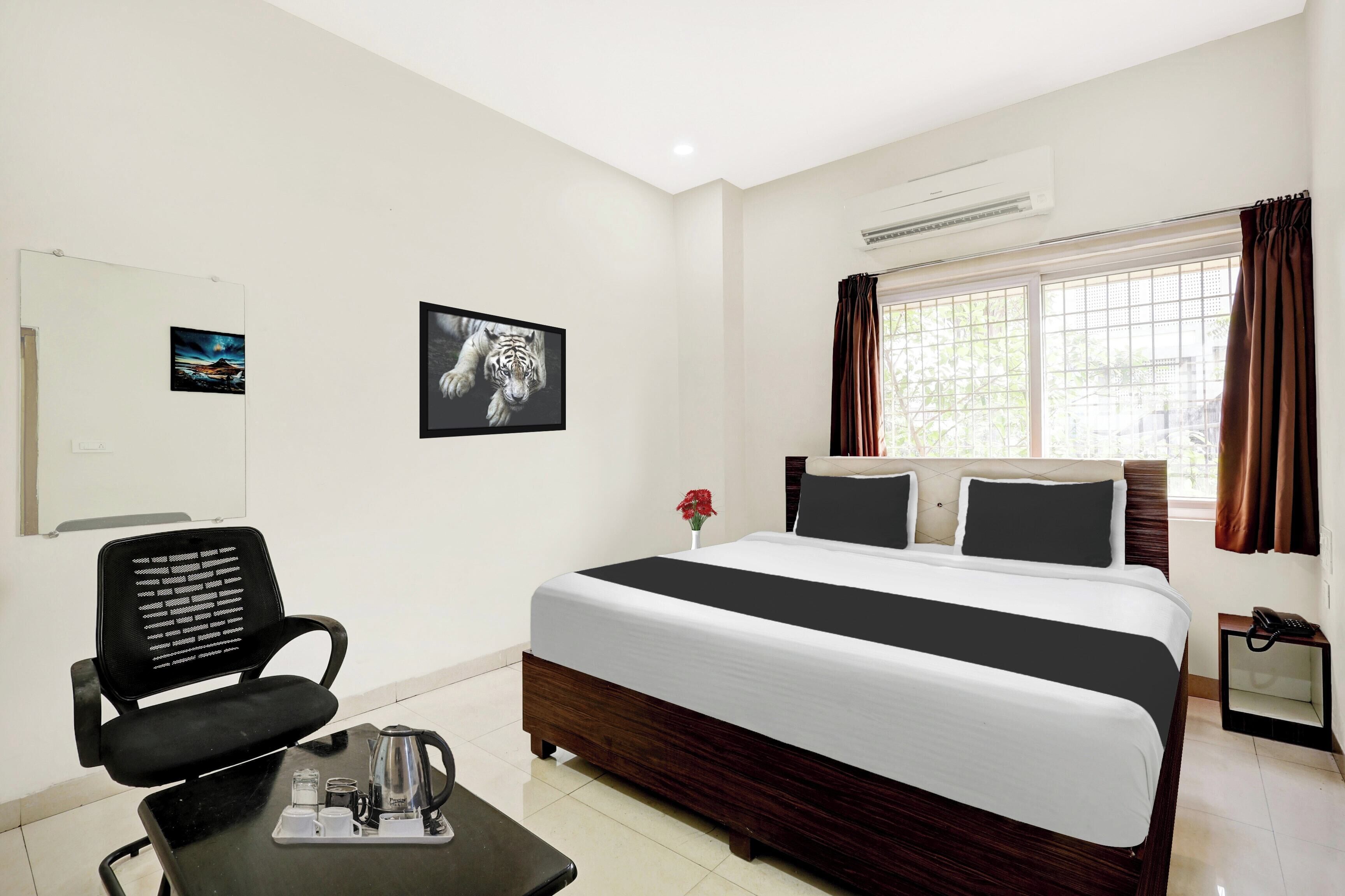 A hotel room with a bed, a chair, a small table with a coffee pot, a picture frame, and a window with curtains.