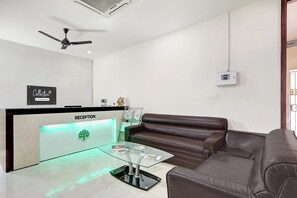 Un espace d’accueil comprenant un comptoir de réception, un ventilateur de plafond, un canapé en cuir marron, une table basse en verre et un thermostat fixé au mur.