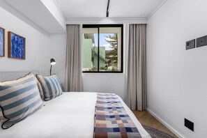 Quarto de hotel com cama, janela com cortinas, abajur e uma obra de arte na parede.