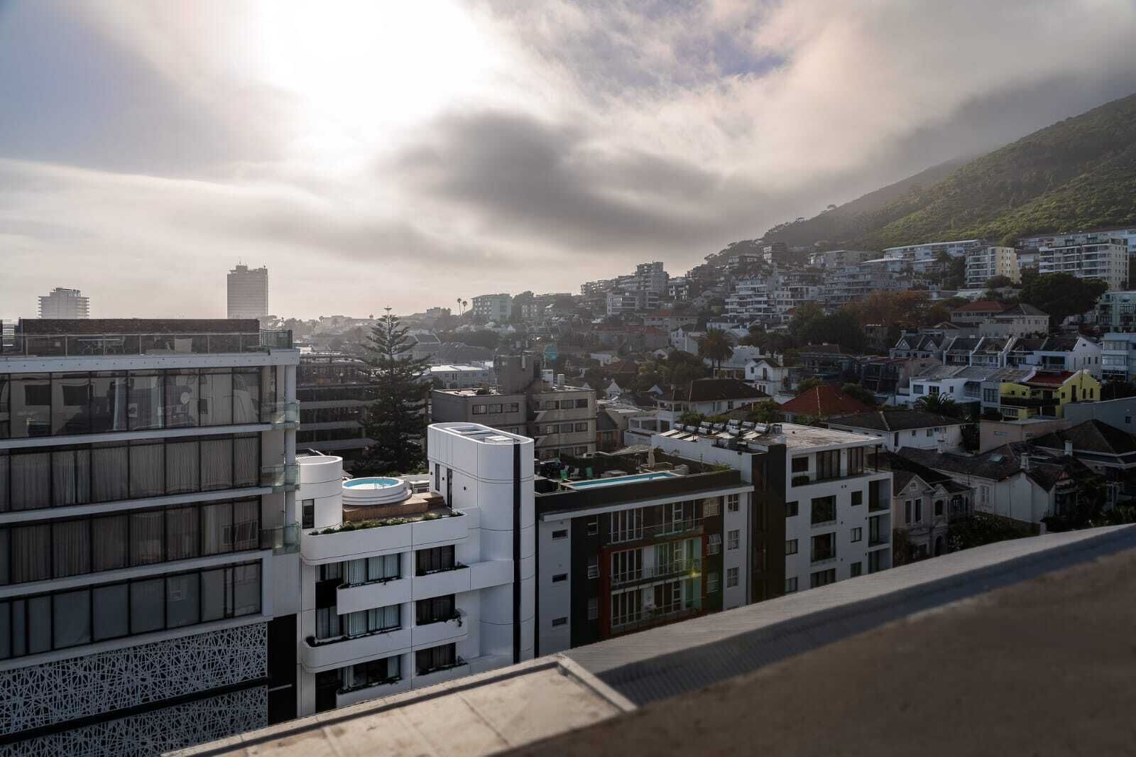 A cityscape with modern buildings, a pool on a rooftop, and a hillside in the background.