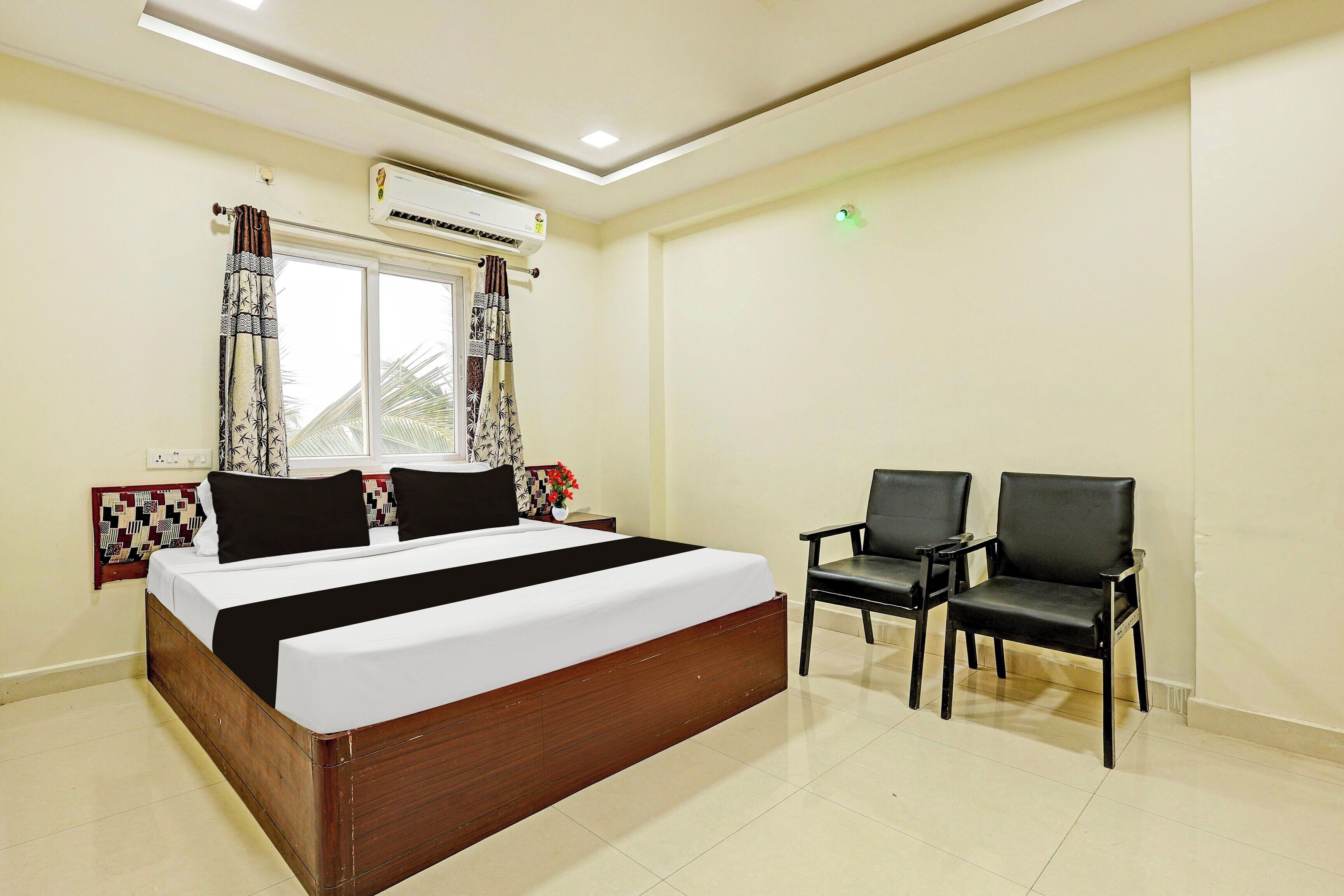 Une chambre d’hôtel avec un grand lit, deux chaises noires, un appareil de climatisation et une fenêtre avec des rideaux à motifs.