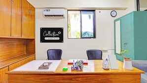 A room with a wooden desk, two chairs, an air conditioning unit, a window with a view, a clock, and a sign indicating the room is company-serviced. - Super Collection O Beach Road Near GITAM (Visakhapatnam)