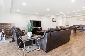 A modern living room with a leather sofa, a glass coffee table, a flat-screen TV, and a brick fireplace. - Greystone Lavender Farm-Hot Tub|Pool Table|KoiPond (Meaford)
