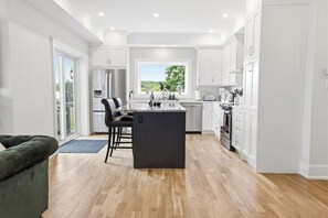 A modern kitchen with a central island, white cabinetry, and a view through a window. - Greystone Lavender Farm-Hot Tub|Pool Table|KoiPond (Meaford)