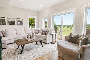A spacious living room with a sofa set, a coffee table, and a view of a green landscape through large windows.