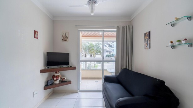 A living room with a black sofa, a TV on a wooden shelf, a ceiling fan, and a view of a balcony with outdoor furniture. - Apartment With Balcony and Barbecue Cac0109 (Florianópolis)