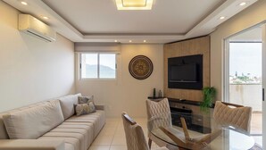 A modern living room with a sofa, a glass dining table, and chairs. There is a flat-screen TV mounted on the wall and a window with a view of the outdoors. - Apart w/ Gourmet Area 240m From the Beach Cac0301 (Florianópolis)