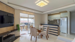 A modern living room with a flat-screen TV, a dining area with a glass table, and a kitchen with a refrigerator and cabinets. - Apart w/ Gourmet Area 240m From the Beach Cac0301 (Florianópolis)