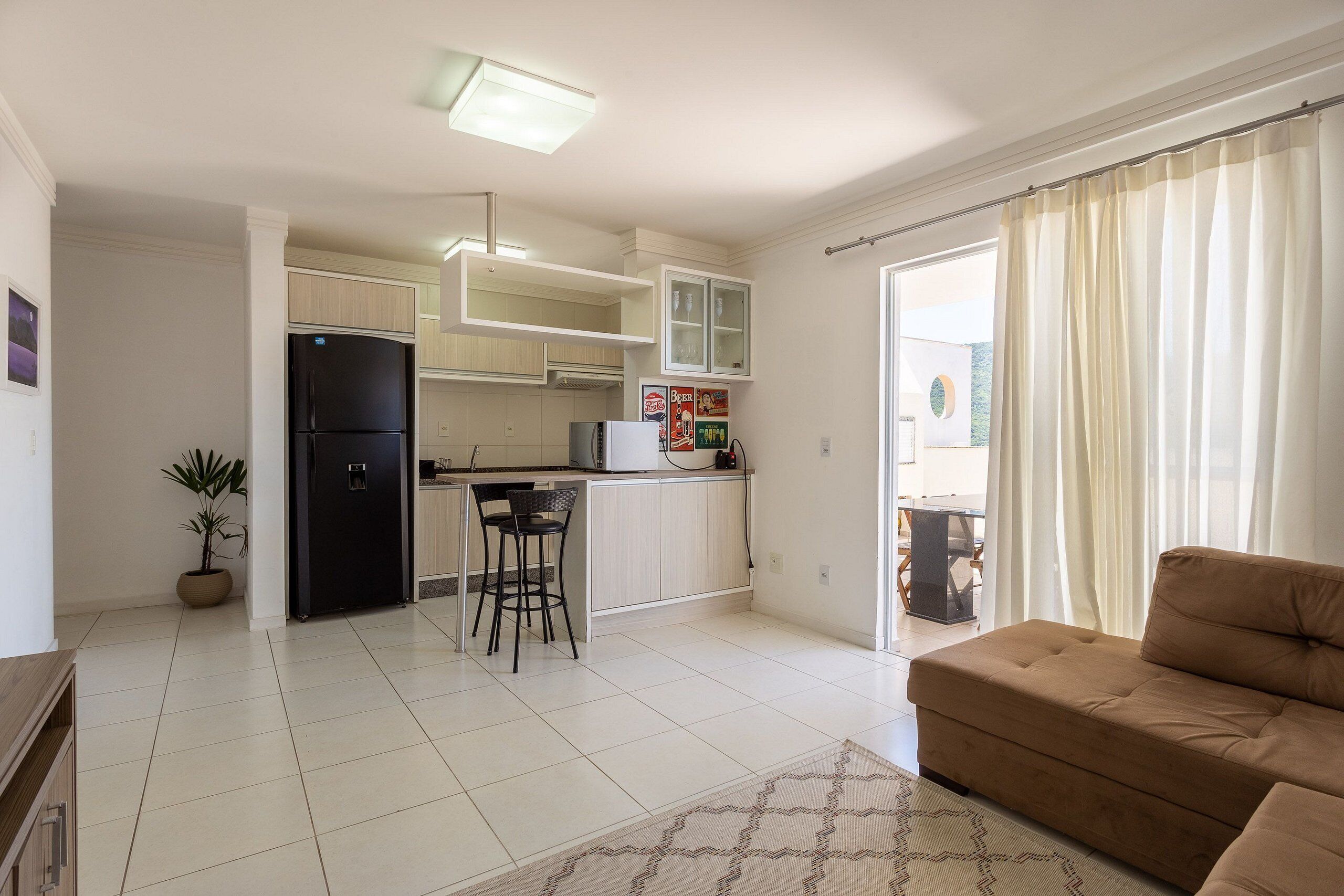 Una cocina moderna con refrigerador, taburetes de bar y una mesa de comedor. Hay un sofá y una planta en maceta en la sala de estar.