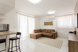 Uma sala de estar moderna com sofá seccional, uma mesa de jantar com cadeiras, área de cozinha com micro-ondas e uma grande janela com cortinas.