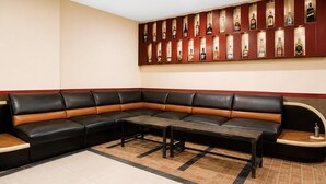 A corner seating area with a long black and tan sofa, a small table, and a wall-mounted liquor display. - Hotel O Kosta Living - Standard Single (Palembang)