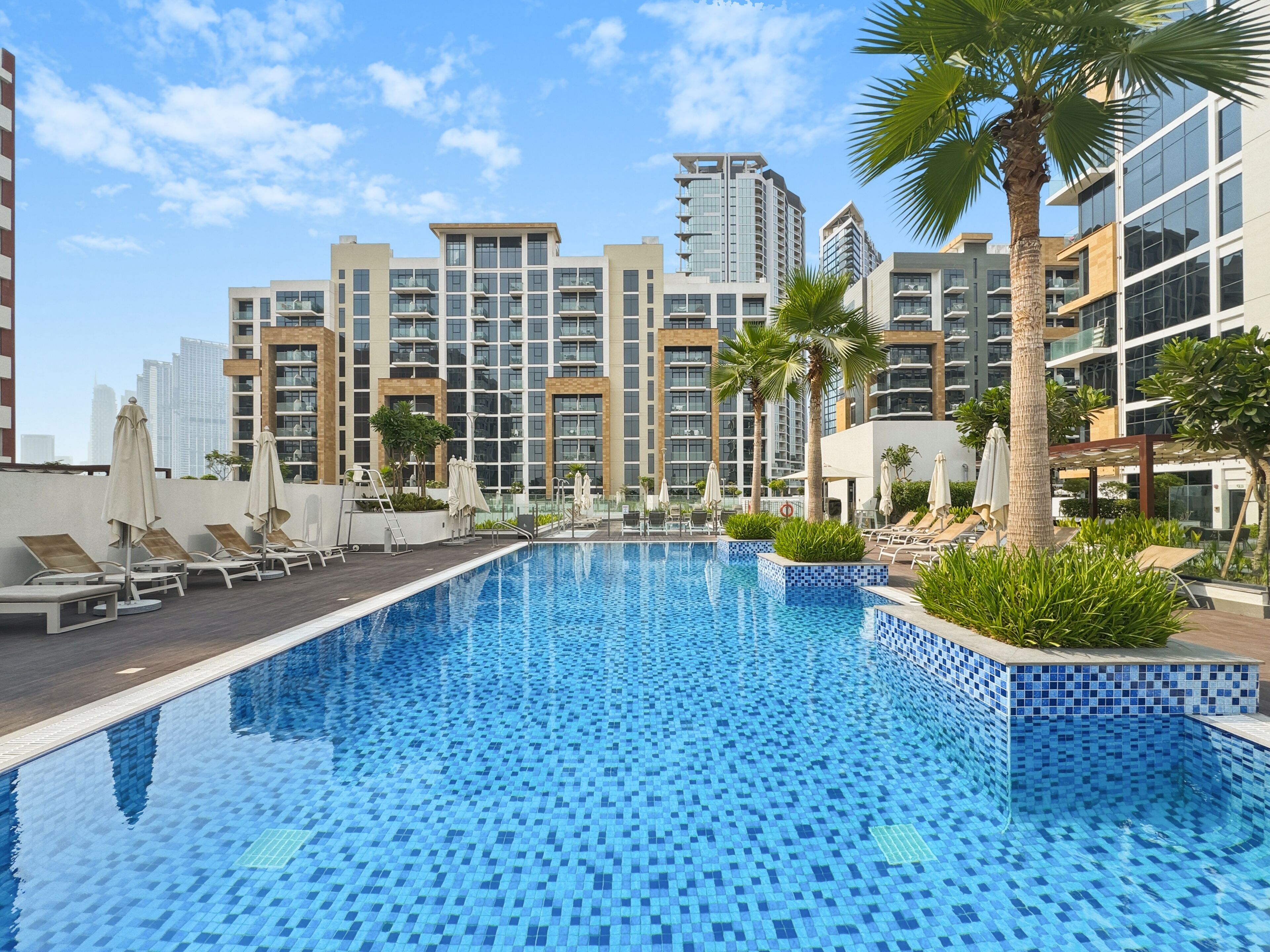 A swimming pool with lounge chairs and umbrellas, surrounded by modern buildings and palm trees.