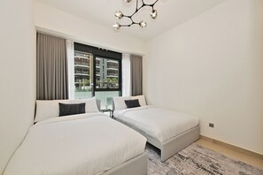 A hotel room with two beds, a large window with curtains, a modern chandelier, and a view of a building outside. - Bloomfields 2BR in Azizi Riviera 43 - Meydan (Dubai)