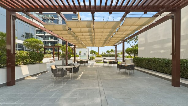 An outdoor patio area with tables and chairs, a pergola structure, and a view of modern buildings in the background. - Bloomfields 2BR in Azizi Riviera 43 (Dubai)