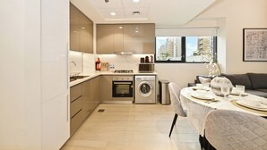 A modern kitchen with built-in appliances, a dining area with a table set for four, and a view of a residential building outside. - Bloomfields 2BR in Azizi Riviera 43 (Dubai)
