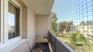 Un balcón con una silla de madera, una mesita con un frutero, y vista a una zona residencial a través de una malla metálica.