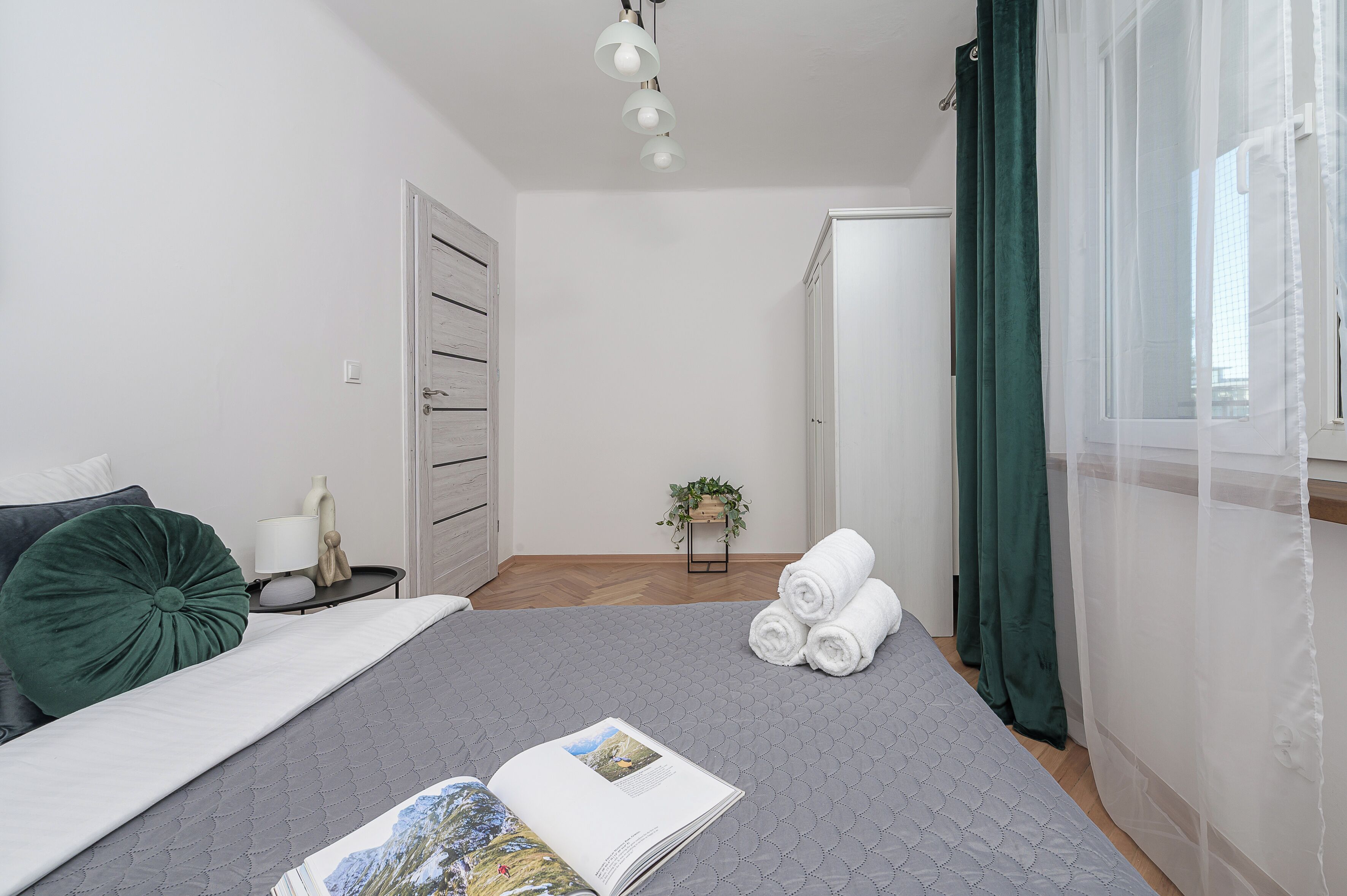 Un dormitorio ordenado con cama, mesita de noche, lámpara, almohada verde, toalla enrollada y un libro sobre la cama.
