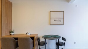 Ein schlicht eingerichteter Raum mit einem Holztisch, zwei Stühlen, einem kleinen Tisch mit einer grünen Unterlage und einem gerahmten Bild an der Wand.