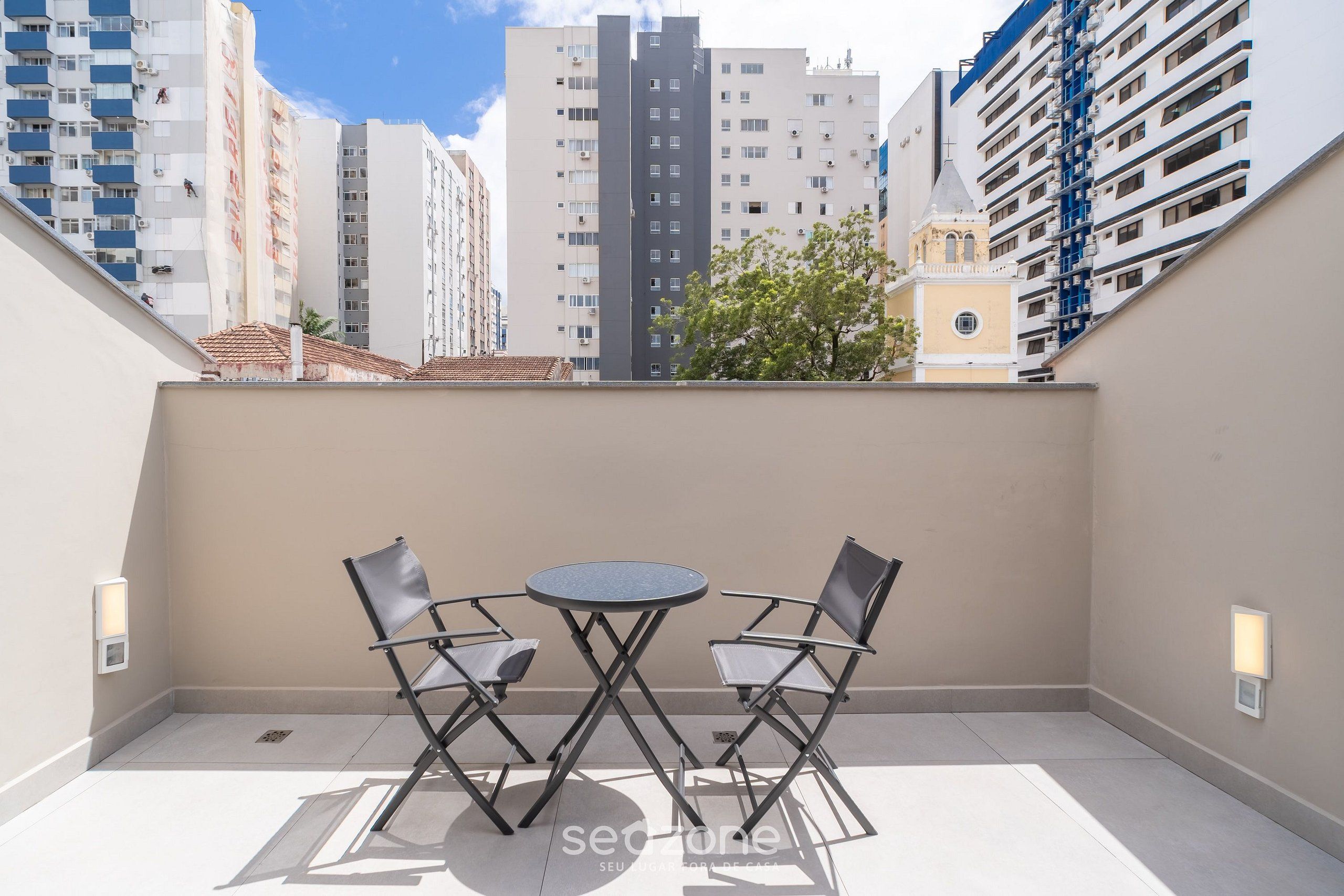 A balcony with two chairs and a small table, overlooking a cityscape with tall buildings.