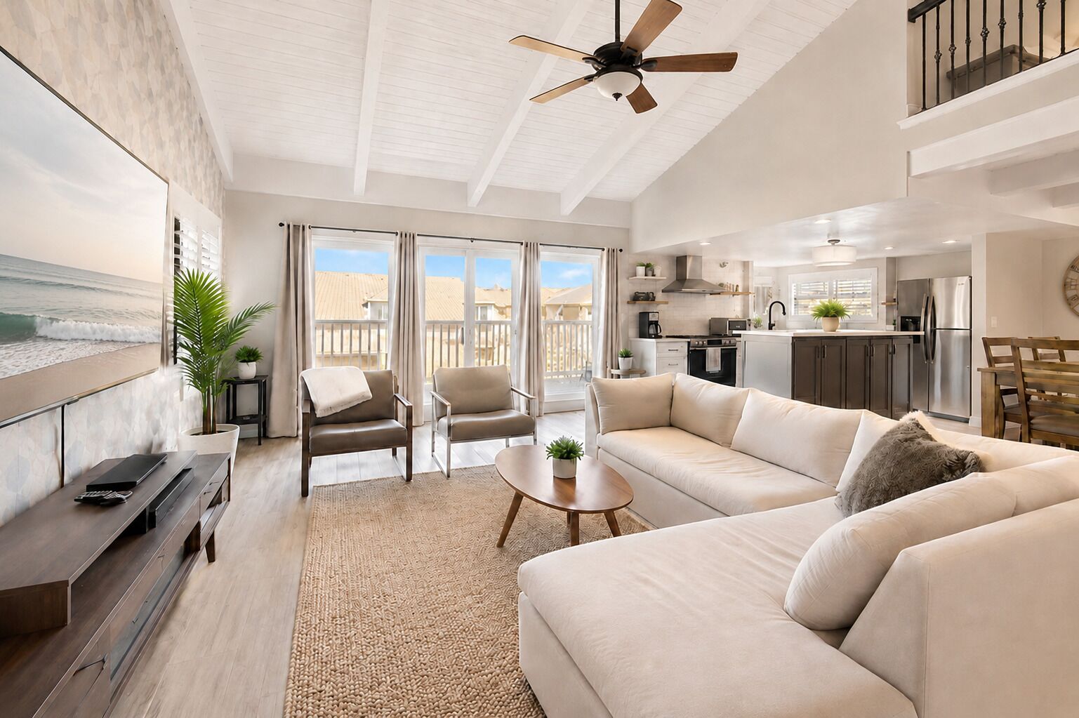 A modern living room with a sofa, chairs, a coffee table, and a TV. There is a balcony with a view of the beach.
