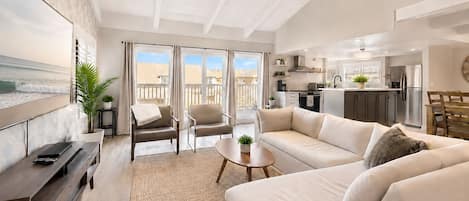 A modern living room with a sofa, chairs, a coffee table, and a TV. There is a balcony with a view of the beach.
