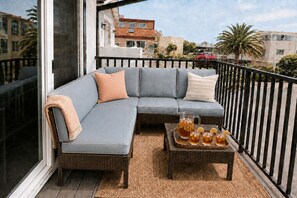 Ein Balkon mit einem grauen Ecksofa, einem Korbtisch mit Getränken und Blick auf Gebäude und Palmen.
