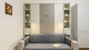 Een kamer met een grijze bank, een boekenkast en een deur.