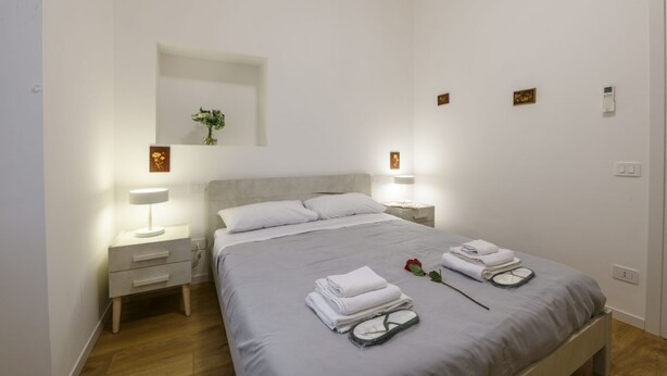 Een opgemaakt bed met gevouwen handdoeken en een klok, een nachtkastje met een lamp en een spiegel aan de muur.