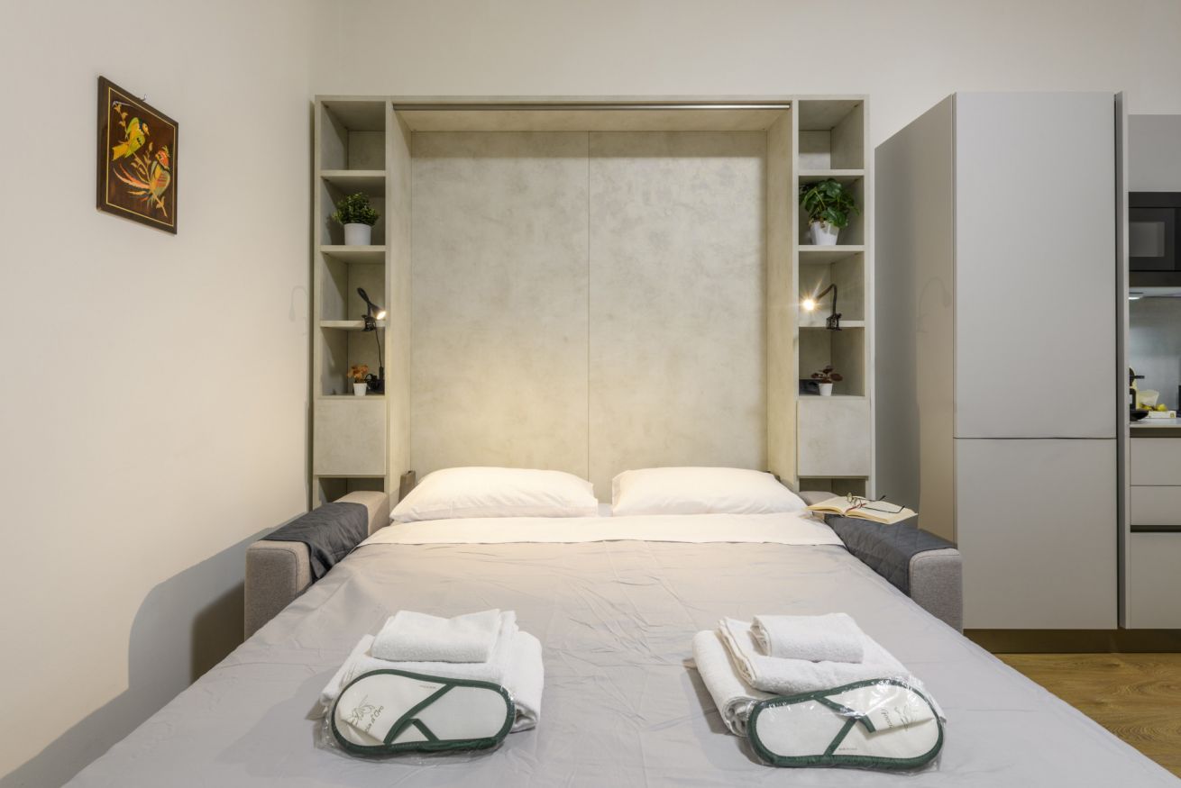 A hotel room with a bed, a bedside table, a wardrobe, and a shelf with plants.
