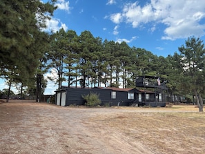 A two-story cabin with a metal exterior, surrounded by tall pine trees, and a dirt ground. - Gated property - 3 bedroom 2 bath plenty of room for your toys (Kingston)