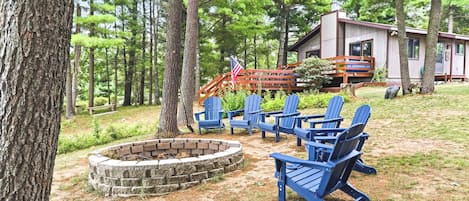 A cabin with a fire pit and blue Adirondack chairs in a wooded area.
