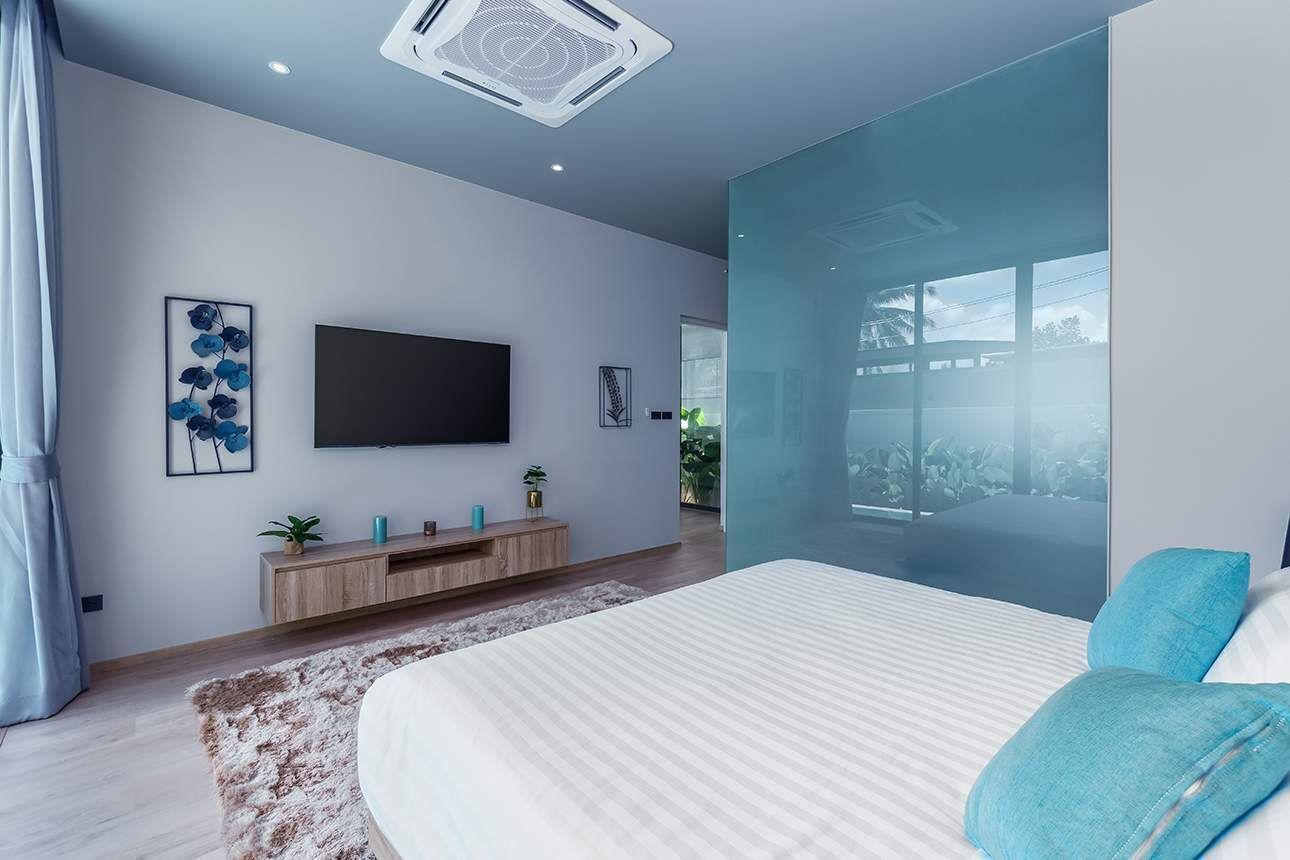 Ein modernes Schlafzimmer mit einem großen Bett, einem flachbildfernseher an der Wand, einem Holztischchen mit Dekorationsgegenständen und einem großen Fenster mit Blick ins Freie.