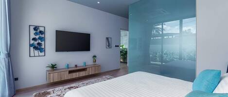 A modern bedroom with a large bed, a flat-screen TV mounted on the wall, a wooden console table with decorative items, and a large window offering a view of the outdoors.