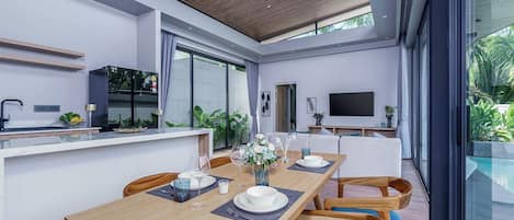 A modern dining area with a wooden table set for four, a kitchen area with a TV, and a large window overlooking a pool.
