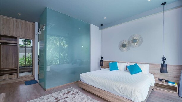 Un dormitorio moderno con un separador de vidrio, una cama grande con ropa de cama blanca y almohadas azules, y un cabecero de madera.