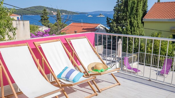 Een balkon met twee houten lounge fauteuils, waarvan er een met een strohoed en kussen, en een uitzicht op zee en heuvels op de achtergrond.