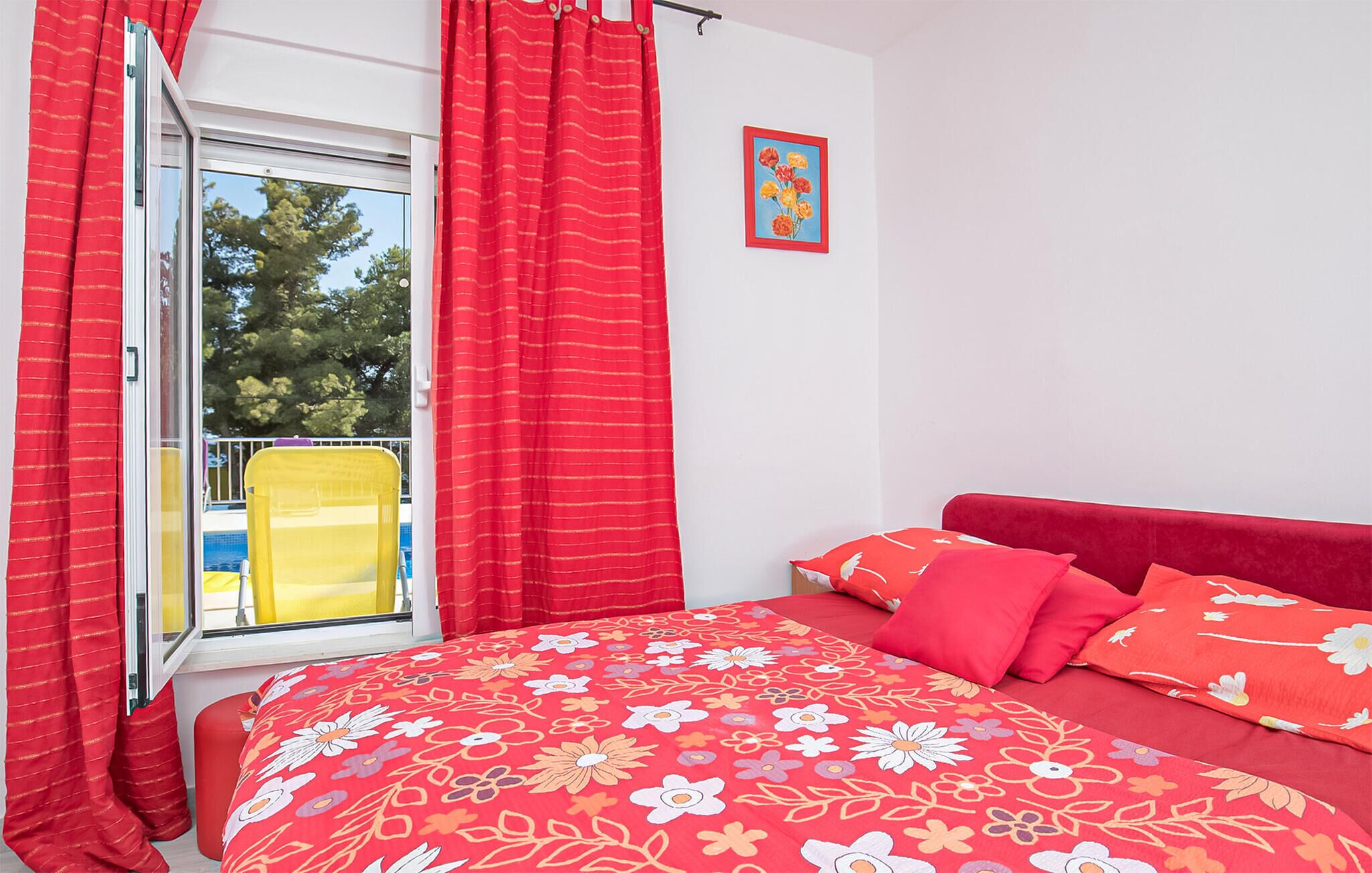 A bedroom with a bed, red curtains, a window, and a yellow chair outside.