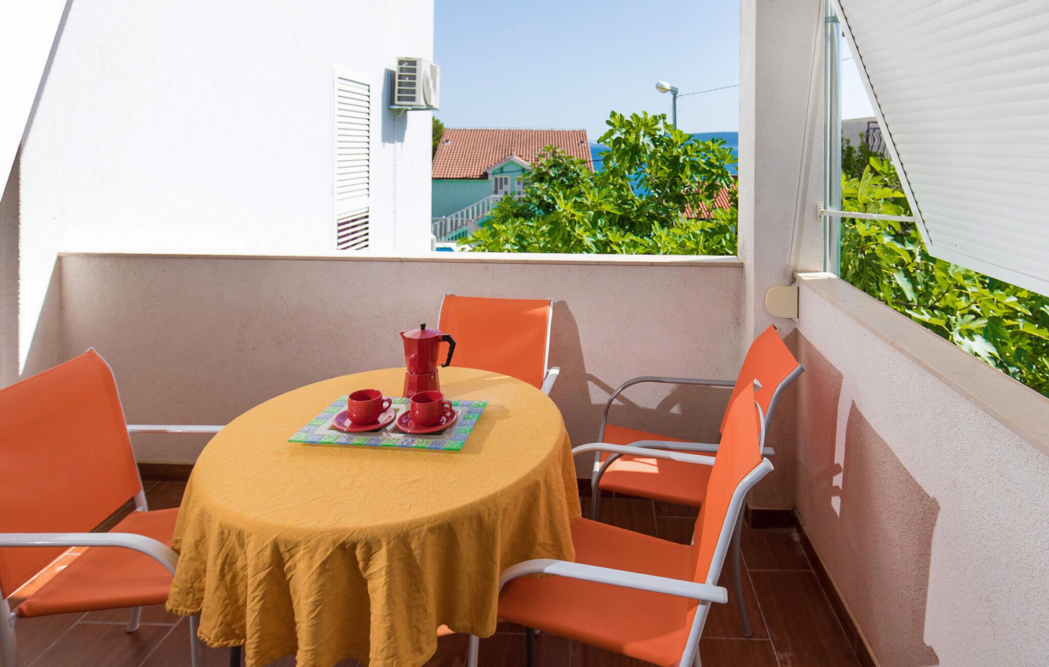 A round table with a yellow tablecloth, four orange chairs, and a red coffee pot on a balcony with a view of a house and greenery.
