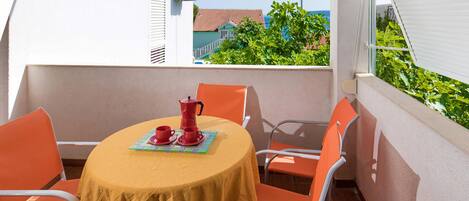 A round table with a yellow tablecloth, four orange chairs, and a red coffee pot on a balcony with a view of a house and greenery.