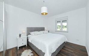 Ein Schlafzimmer mit einem Bett, Nachttischen, einer Hängeleuchte, einem Fenster und Blick ins Grüne.