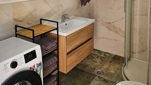 A compact bathroom with a washing machine, a wooden shelf with towels, a sink, and a shower area. - Awesome home in Ervenik (Ervenik)