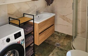A compact bathroom with a washing machine, a wooden shelf with towels, a sink, and a shower area. - Awesome home in Ervenik (Ervenik)