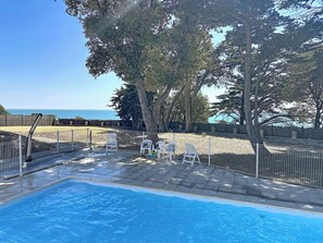 A swimming pool with a view of the sea, surrounded by a fenced patio area with chairs and a wooden fence. - Les Sylphes by Interhome (Pornic)