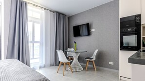 A modern hotel room with a bed, a dining table with chairs, a flat-screen TV mounted on the wall, and a kitchenette with an oven.