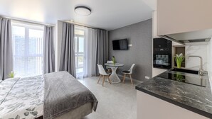 A modern hotel room with a bed, a dining area with chairs, a TV mounted on the wall, and a kitchen area with a sink and stove.