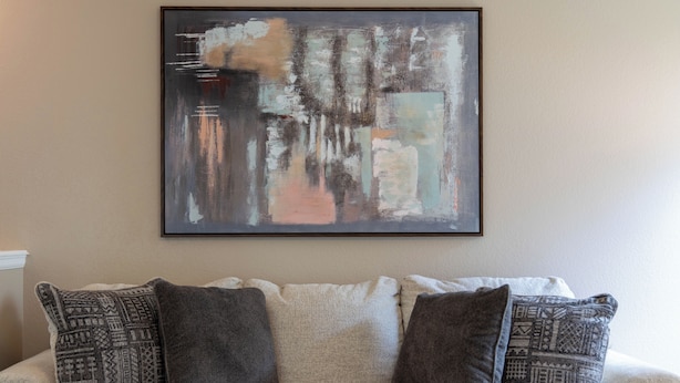 A living room with a beige sofa, three patterned cushions, and a large abstract painting on the wall.